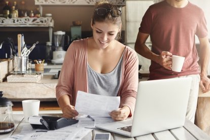 Young pretty woman wearing glasses on her head smiling happily while reading document saying that bank approved their mortgage application, her husband standing on background and drinking coffee