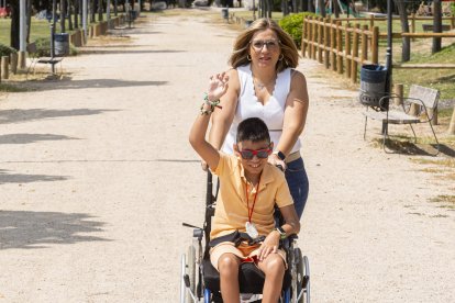 Álex saluda con las pulseras que ha hecho en el campamento y luce su colgante de arcilla, empujado en silla de ruedas por su madre, Leticia Merino.- PHOTOGENIC