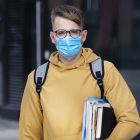 Guy student, pupil boy, young man in protective medical mask and glasses on face outdoors university with books, textbooks look at camera. Virus, pandemic coronavirus concept. Covid-19, safety study