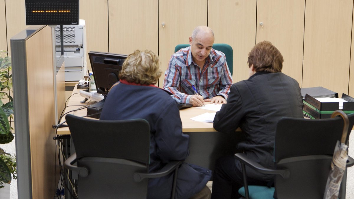 Un empleado público atiende a dos personas en una oficina de atención ciudadana.
